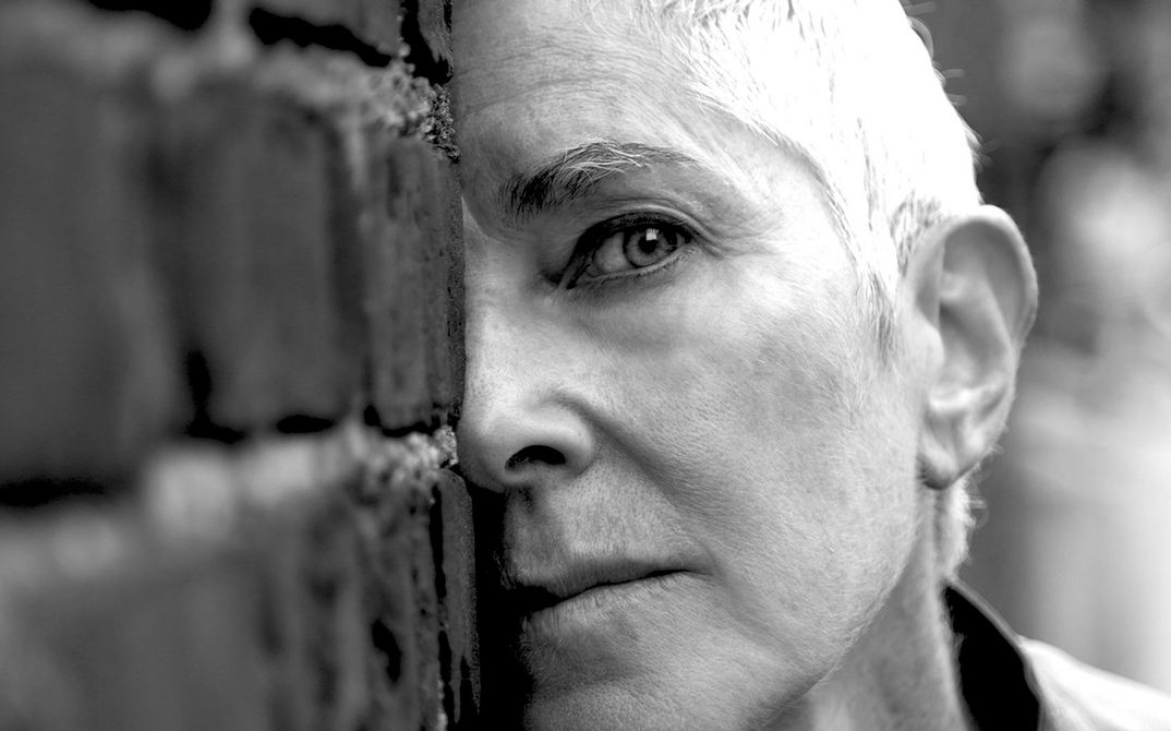 Portrait photo of Beth B. She wears her hair short and looks directly into the camera. Parts of her face are hidden behind a wall.