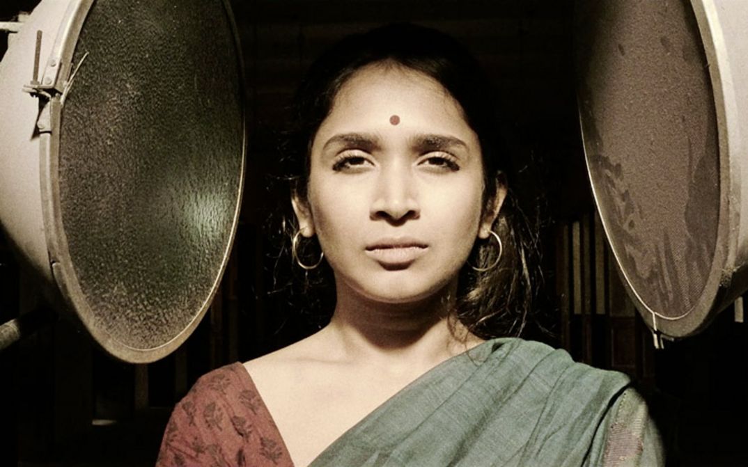 Still from the film "Those Who Do Not Drown" by Naeem Mohaiemen. A woman looks into the camera, two unlit lamps are pointed at her.