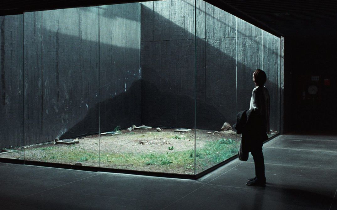Film still from MEMORIA: A woman looks at a glass box. Light falls in through a light shaft.