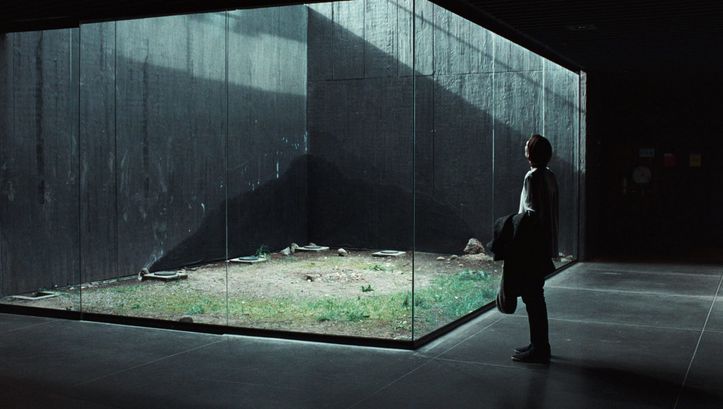 Film still from MEMORIA: A woman looks at a glass box. Light falls in through a light shaft.
