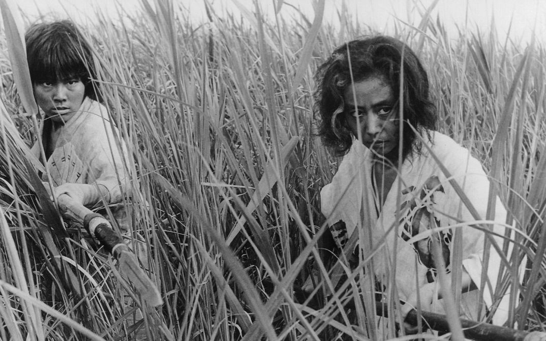 Film still from ONIBABA: Two women, armed with spears, stand in an attack position in a field full of reeds.