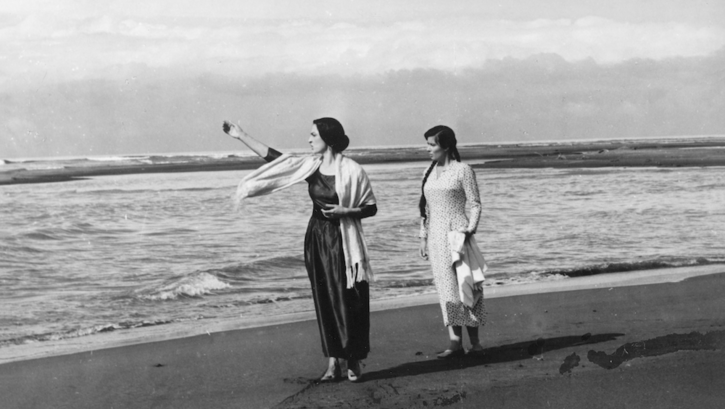 Film still from UNDER ONE SKY: Two young women walk barefoot along a beach.