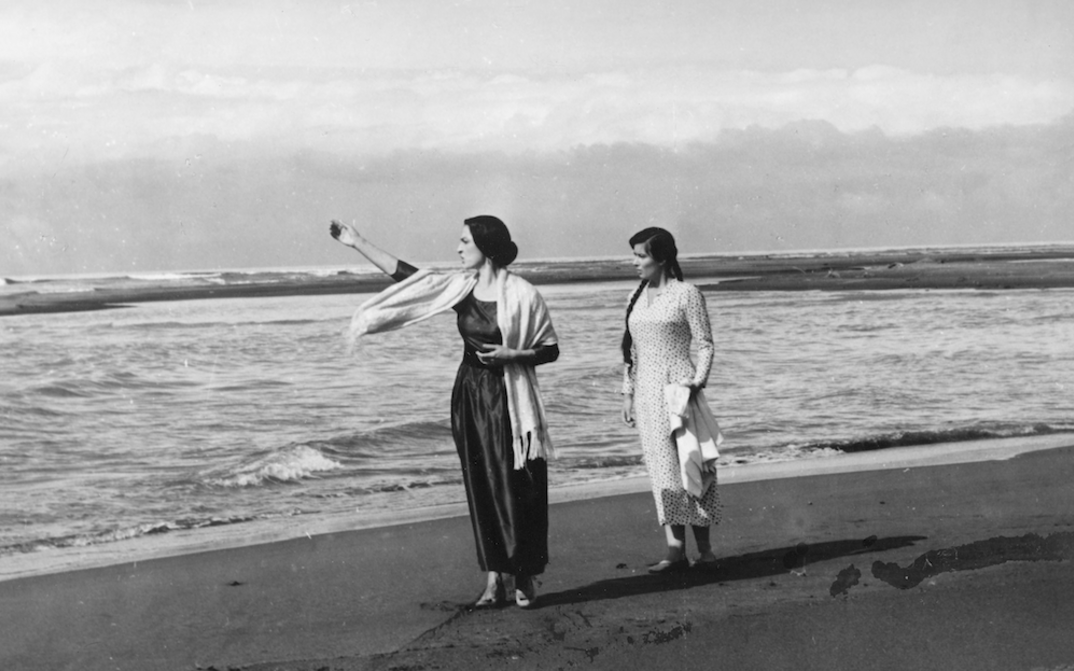 Film still from UNDER ONE SKY: Two young women walk barefoot along a beach.