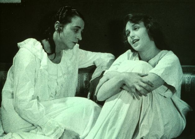 Monochrom eingefärbtes Filmstill aus NORRTULLSLIGAN: Zwei jungen Frauen in Nachthemden sitzen zusammen auf einem Sofa und unterhalten sich.