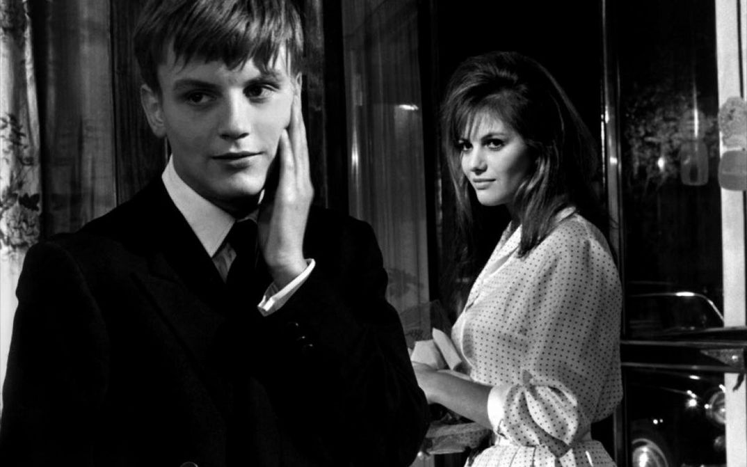 Film still from LA RAGAZZA CON LA VALIGIA: A young man in a suit stands in the foreground of the picture, touching his cheek. Behind him stands a young woman, looking at him.