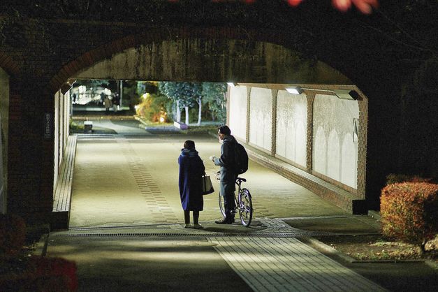 Film still from "Yoake no subete" by Shô Miyake. It shows two people talking under a pedestrian tunnel. The person on the right is carrying a bicycle. It is dark, the tunnel is illuminated. 
