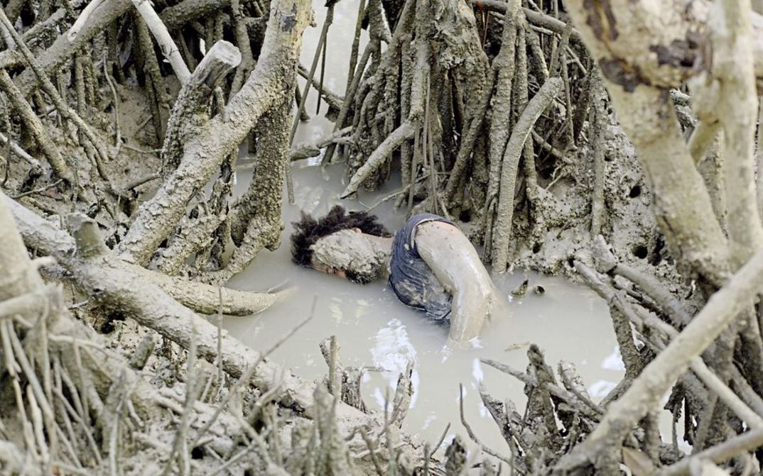 Filmstill aus "Resonance Spiral" von Filipa César und Marinho de Pina. Zu sehen ist eine mit Schlamm bedeckte Person in einem Teich voller Baumwurzeln und Äste. 