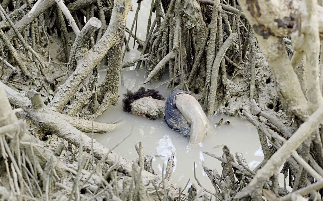 Filmstill aus "Resonance Spiral" von Filipa César und Marinho de Pina. Zu sehen ist eine mit Schlamm bedeckte Person in einem Teich voller Baumwurzeln und Äste.