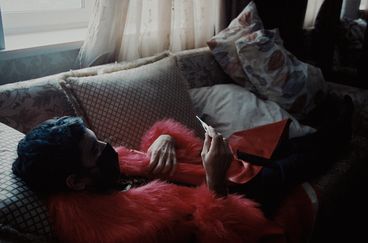 A man wearing black face mask and a red fur and leather jacket lays on a couch inspecting the results of a COVID test.