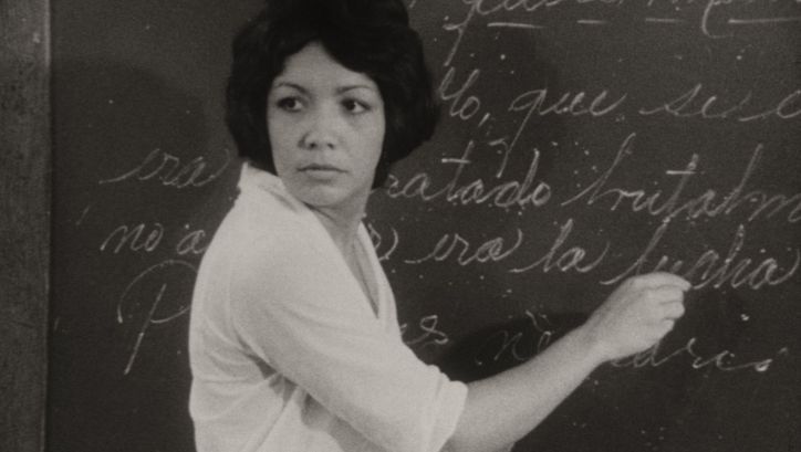 Filmstill aus DE CIERTA MANERA: Eine Frau schreibt in einem Klassenzimmer an die Wandtafel.