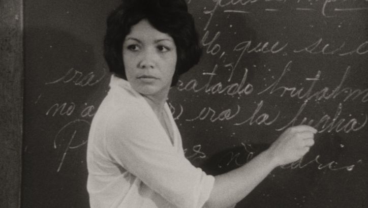 Film still from DE CIERTA MANERA: A woman writes on the blackboard in a classroom.