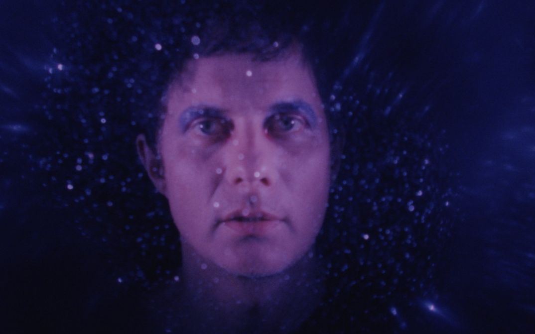 Filmstill aus FELIX IN WONDERLAND: Vor einem dunklen Hintergrund sieht man das Gesicht eines Mannes. Er blickt direkt in die Kamera, trägt die Haare kurz und blaues Augen-Make-up.