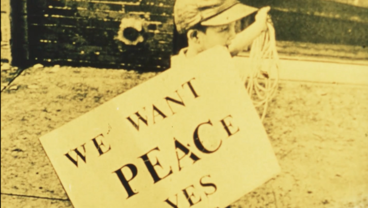 Film still from FOR LIFE, AGAINST WAR: A boy carries a sign saying "We want peace yes".