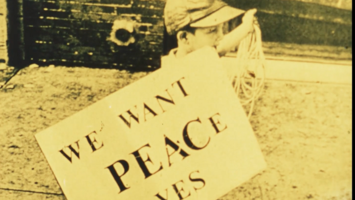 Filmstill aus FOR LIFE, AGAINST WAR: Ein Junge trägt ein Schild mit dem Aufdruck "We want peace yes".