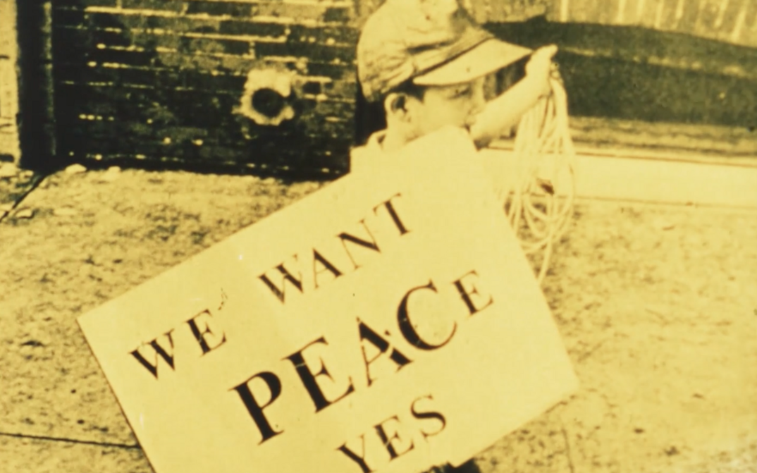 Filmstill aus FOR LIFE, AGAINST WAR: Ein Junge trägt ein Schild mit dem Aufdruck "We want peace yes".