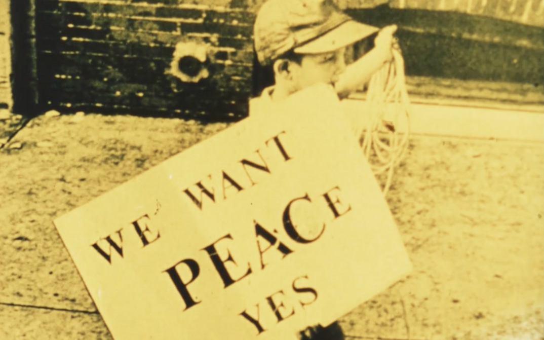 Film still from FOR LIFE, AGAINST WAR: A boy carries a sign saying "We want peace yes".