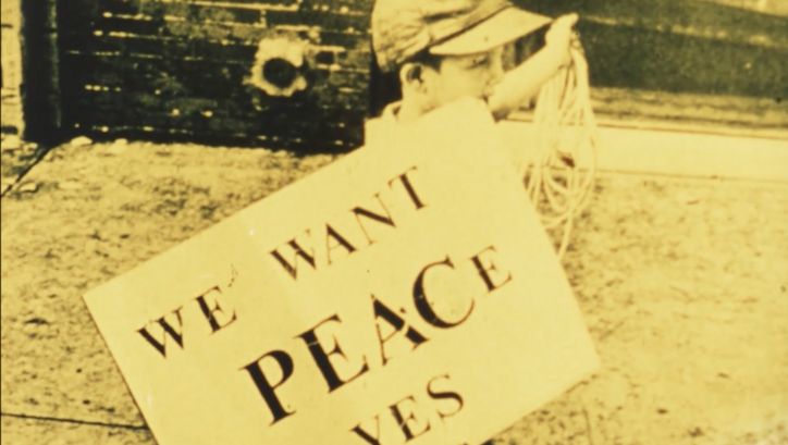 Film still from FOR LIFE, AGAINST WAR: A boy carries a sign saying "We want peace yes".