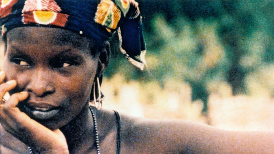 Filmstill aus NAKED SPACES – LIVING IS ROUND: Eine Frau mit einem bunten Tuch auf dem Kopf blickt nachdenklich in die Ferne und hat den Kopf in eine Hand gestützt.