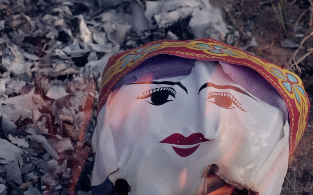 Still from the film "Memoryland" by Kim Quy Bui. We see a plastic ceremonial face engulfed in flames.