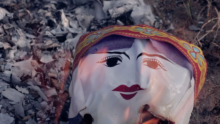 Still from the film "Memoryland" by Kim Quy Bui. We see a plastic ceremonial face engulfed in flames. 