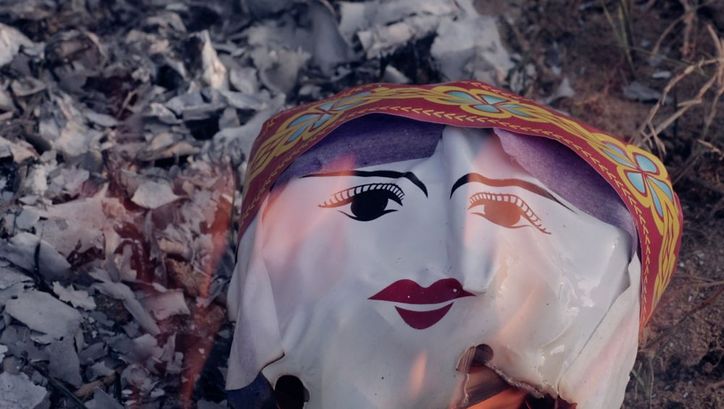 Still from the film "Memoryland" by Kim Quy Bui. We see a plastic ceremonial face engulfed in flames. 
