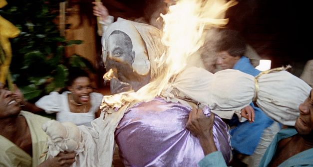 Filmstill aus „West Indies" von Med Hondo. Eine Gruppe von lachenden aufgeregten Menschen hebt eine brennende lebensgroße Lumpenpuppe hoch. 