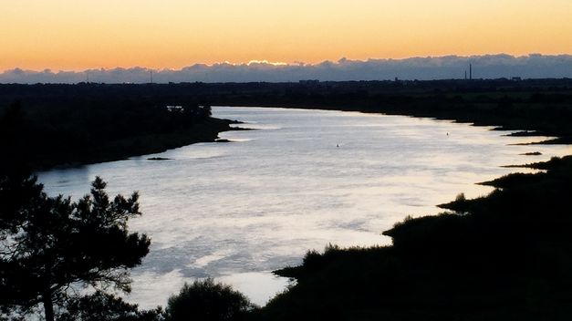 Landschaftsaufnahme eines breiten Flusses bei Sonnenuntergang, Ufer und Bäume liegen im Schatten.