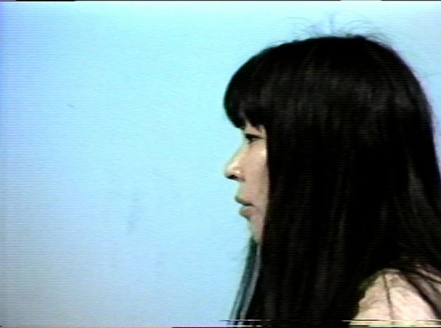Still from “I Am (Not) Takahiko Iimura, I Am (Not) Akiko Iimura”: a woman in profile with long black hair against a blue background (the artist’s wife).