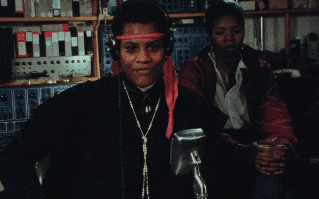 Film still from BORN IN FLAMES: Two women in a radio studio, one standing in front of a microphone with headphones.
