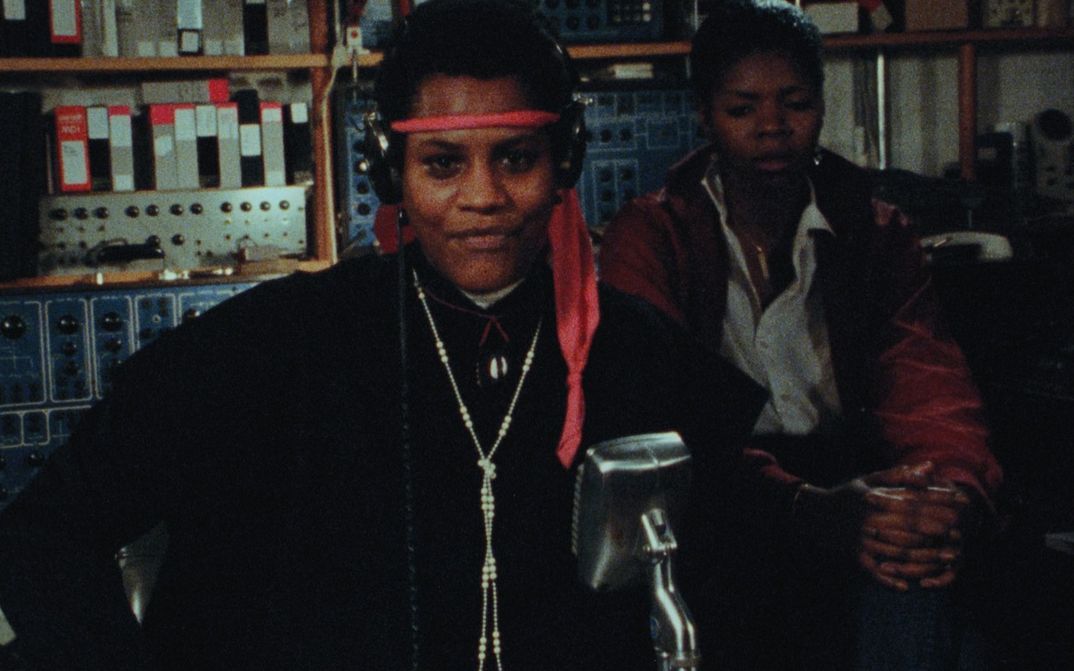 Filmstill aus BORN IN FLAMES: Zwei Frauen in einem Radiostudio, die eine steht mit Kopfhörern vor einem Mikrophon.