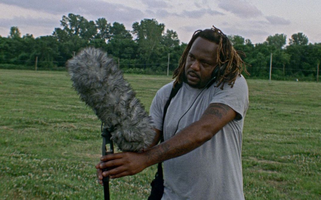Film still from Theresa Delsoin, Lisa Malloy, JP Sniadecki, Ray Whitaker’s film “On the Battlefield”. A sound recordist stands in a field holding a boom pole and wearing headphones, and listens intently to what they are recording. In the background we see a cloudy sky and trees.