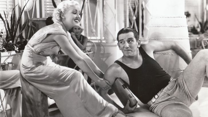 Film still from THE GAY DIVORCEE: Edward Everett Horton is sitting on a sofa in shorts and a tank top, a young woman playfully tugging at him.
