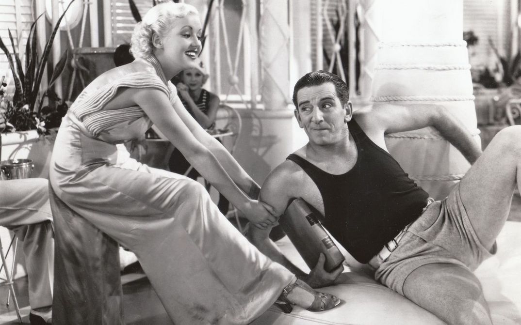 Film still from THE GAY DIVORCEE: Edward Everett Horton is sitting on a sofa in shorts and a tank top, a young woman playfully tugging at him.