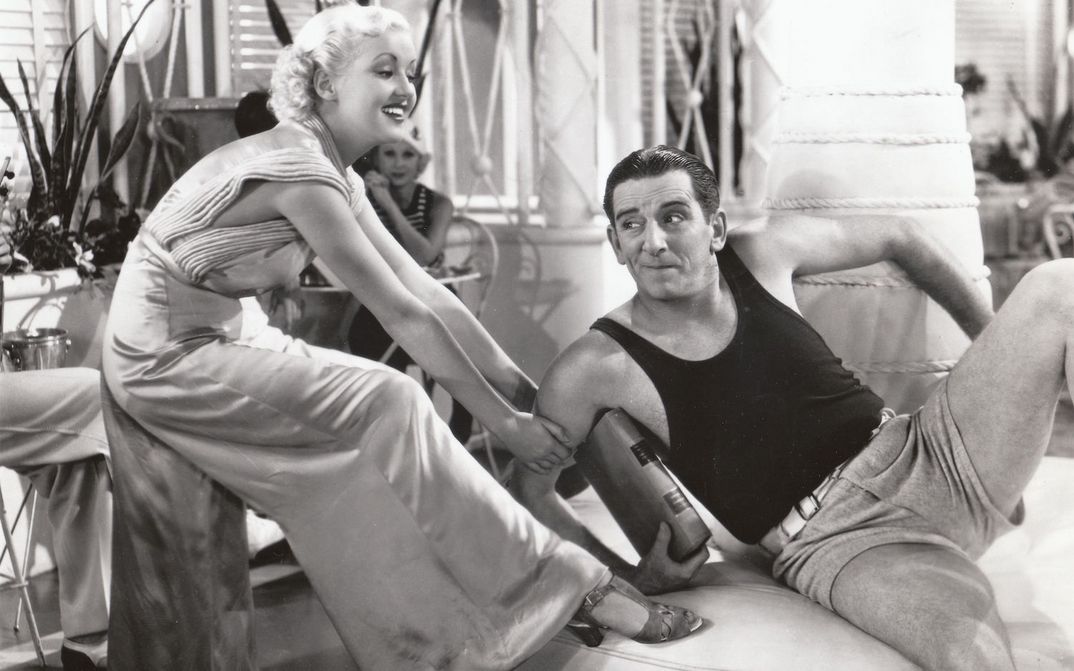 Film still from THE GAY DIVORCEE: Edward Everett Horton is sitting on a sofa in shorts and a tank top, a young woman playfully tugging at him.