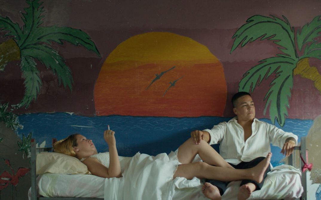 Still aus dem Film REAS von Lola Arias (2023): Eine Frau und eine Mann auf einem Bett. Dahinter ein Wandbild eines Sonnenuntergangs mit Palmen.