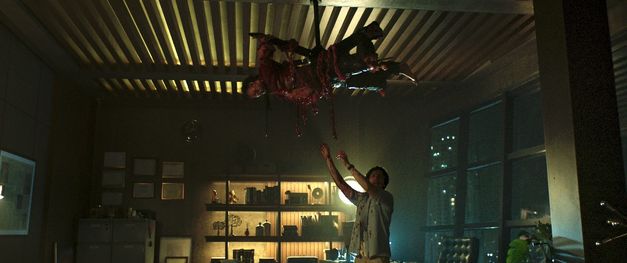 Mann steht in einem Büro. Am Deckenventilator hängt gefesselt eine blutbeschmierte Person.