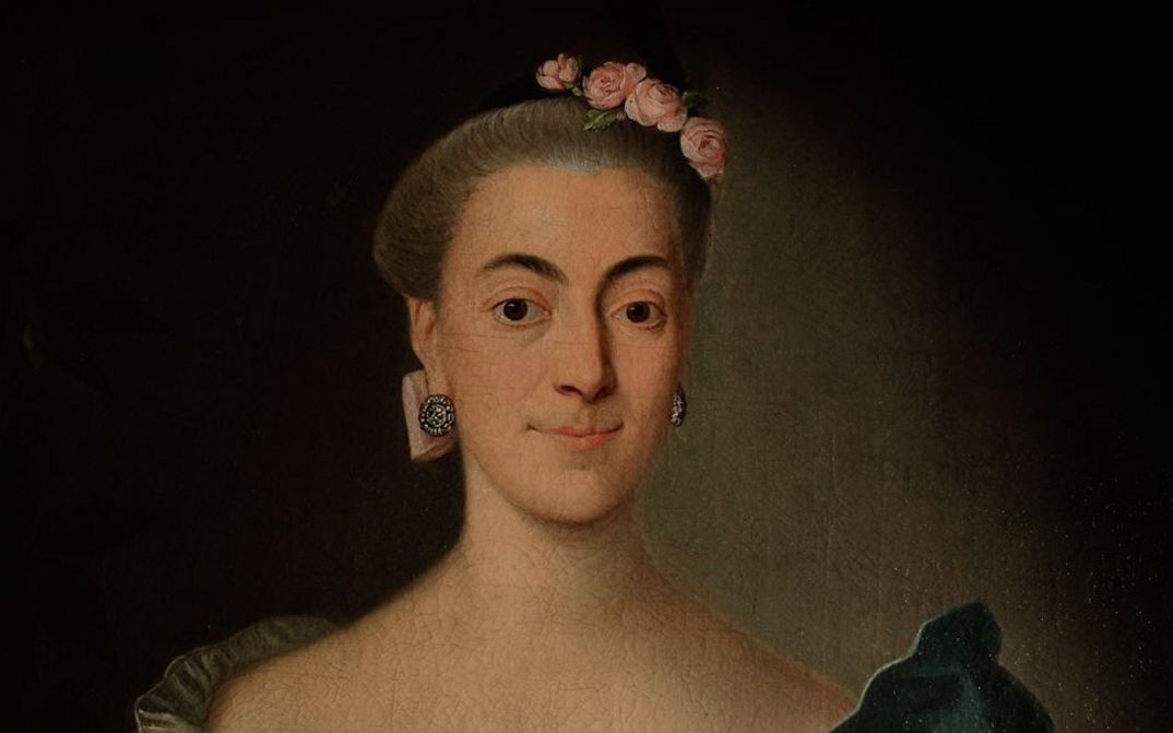 Filmstill aus "Ihre ergebenste Fräulein" von Eva C. Heldmann. Zu sehen ist eine Nahaufnahme des Bildes einer Frau mit hochgestecktem Haar, Blumen auf dem Kopf und runden Ohrringen. 