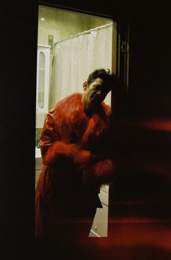 Ein schattiges Bild eines Mannes in einem roten Mantel aus Pelz und Leder, der im Türrahmen eines Badezimmers steht. Er schaut die Kamera an. 