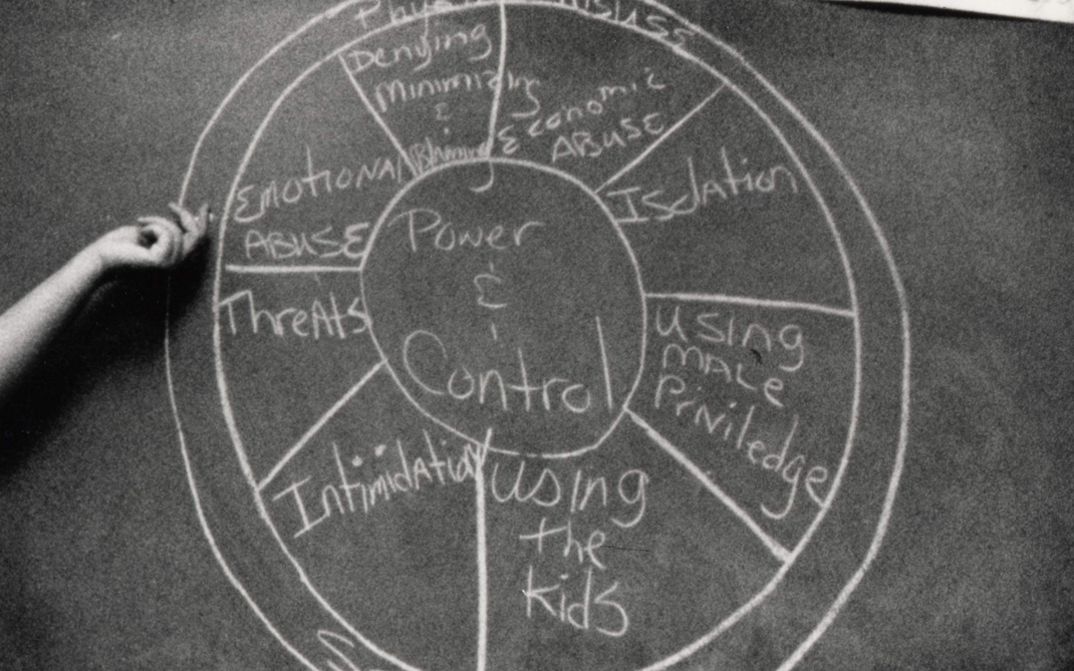 Filmstill aus DOMESTIC VIOLENCE. Auf einer Wandtafel ist mit Kreide ein Diagramm über Methoden häuslicher Gewalt aufgezeichnet.