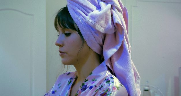 Film still from ENDURANCE: A woman with dark hair in profile. She is wearing a bathrobe and has a towel wrapped around her head.