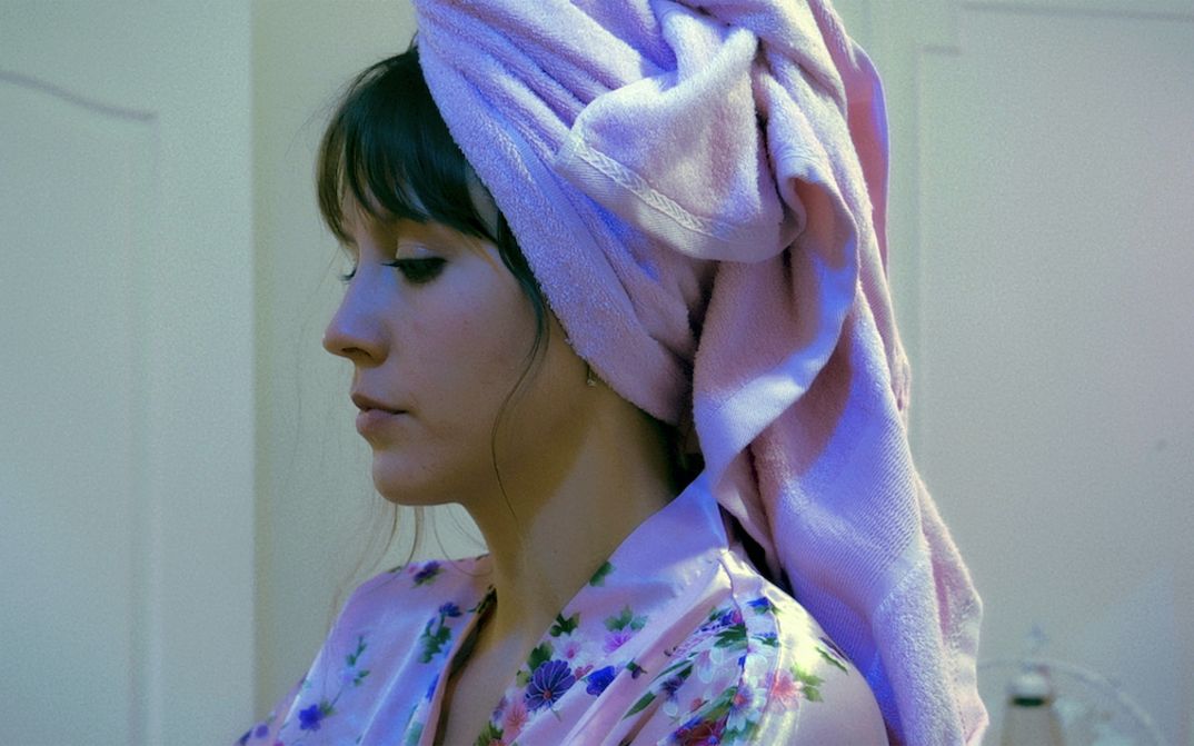 Film still from ENDURANCE: A woman with dark hair in profile. She is wearing a bathrobe and has a towel wrapped around her head.