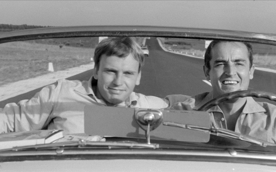 Film still from IL SORPASSO: Two men at the wheel and in the passenger seat of a convertible. The straight and empty road can be seen behind them.