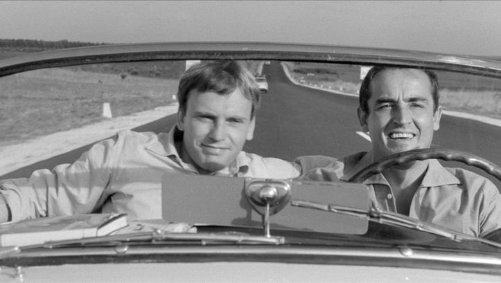 Film still from IL SORPASSO: Two men at the wheel and in the passenger seat of a convertible. The straight and empty road can be seen behind them.