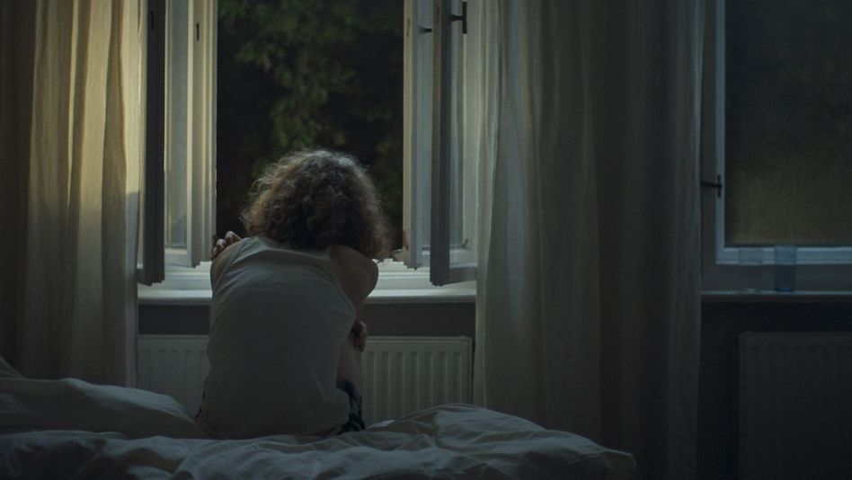 Film still from LANGE GEHST DU VOR MIR HER: From behind, you can see a person with medium-length curly hair sitting in bed at night in front of an open window.