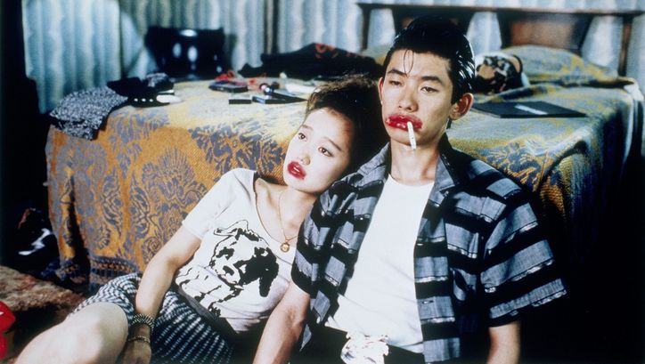 Film still from MYSTERY TRAIN: A man and a woman are sitting on the floor next to a bed. Their mouths are painted a grotesque red, and he has a cigarette in the corner of his mouth.