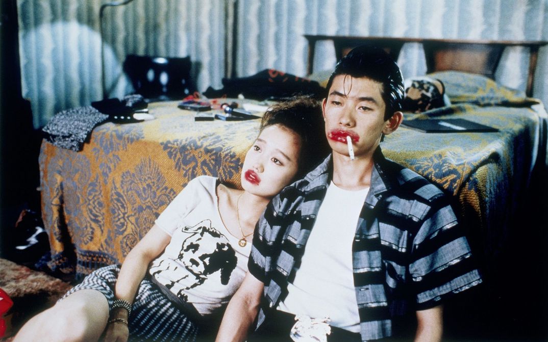 Film still from MYSTERY TRAIN: A man and a woman are sitting on the floor next to a bed. Their mouths are painted a grotesque red, and he has a cigarette in the corner of his mouth.