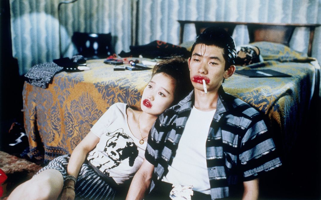 Filmstill aus MYSTERY TRAIN: Ein Mann und eine Frau sitzen an ein Bett gelegt am Boden. Ihre Lippen sind grotesk rot geschminkt, er hat eine Zigarette im Mundwinkel. 