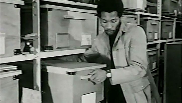 Film still from YOU HIDE ME: A man pulls a large box from a shelf.