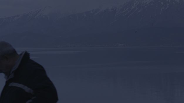 Film still from Deniz Şimşek’s film “detours while speaking of monsters”. A desaturated mountain landscape lies behind a smooth body of water at twilight, and in the foreground a person walks out of the frame left. 