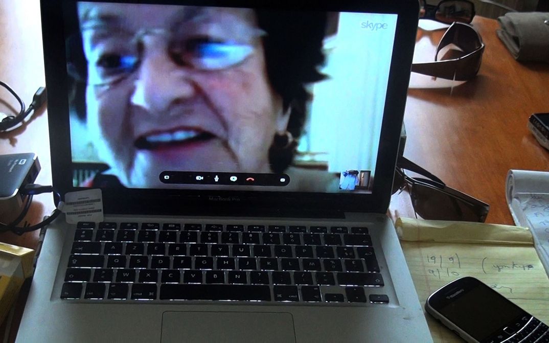 Filmstill aus NO HOME MOVIE: Auf einem Tisch steht ein Laptop, auf dem man eine ältere Frau beim Skype-Gespräch sieht.